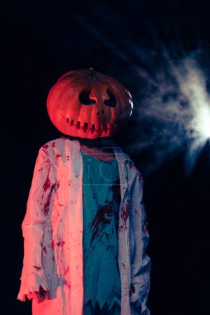 Téléchargez les photos : Jeune femme tient citrouille sur sa tête et dépeint zombie sanguinaire avec des vêtements sanglants sur fond sombre avec contre-jour. Image effrayante pour Halloween. - en image libre de droit