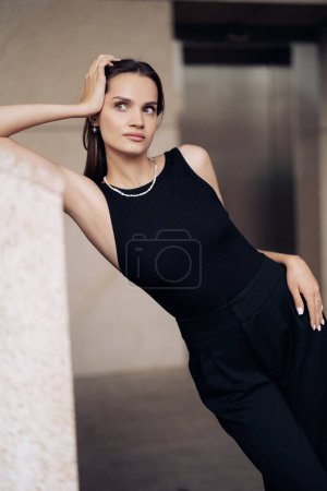 Téléchargez les photos : Jeune jolie femme en robe noire avec collier se tient sur la rue penché près du mur du bâtiment. Gros plan. - en image libre de droit