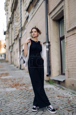 Téléchargez les photos : Jeune jolie femme en robe noire longue et pull drapé sur les épaules se dresse sur la rue près du bâtiment. - en image libre de droit