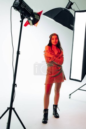 Photo for Young woman poses in studio in knitted lacy dress and bandana near spotlights on tripods and reflector against white background with red backlight. - Royalty Free Image