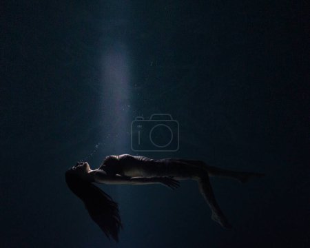 Underwater shoot of beautiful woman swimming and relaxing in water in sunbeams. Fantasy mermaid against water surface background with rays of lights.