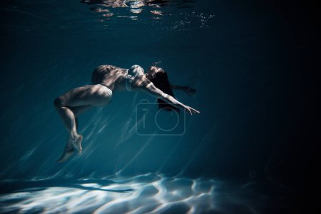 Underwater shoot of beautiful pregnant woman swimming in water through sunbeams. Fantasy mermaid against turquoise water background with rays of lights.
