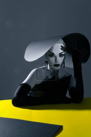 Foto de Una joven con maquillaje en la cara posa en el estudio con un elegante sombrero de ala ancha, vestido negro y guantes. - Imagen libre de derechos