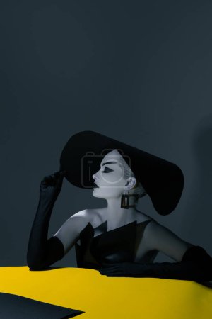 Foto de Una joven con maquillaje en la cara posa en el estudio con un elegante sombrero de ala ancha, vestido negro y guantes. - Imagen libre de derechos