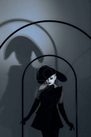 Foto de Mujer joven con maquillaje en la cara posa en el estudio con elegante sombrero de ala ancha, vestido negro y guantes contra su sombra en la pared. - Imagen libre de derechos