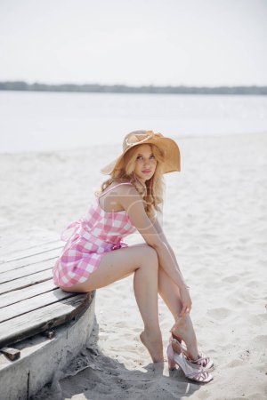 Foto de Joven chica rubia se ven como una muñeca Barbie en rosa mini vestido a cuadros y sombrero de ala ancha sentado descalzo en la playa contra la arena y el fondo de agua. - Imagen libre de derechos