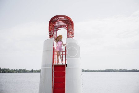 Foto de Joven chica rubia se parece a una muñeca Barbie en rosa mini vestido a cuadros y sombrero de ala ancha de pie en la torre de la playa salvavidas contra el agua y el fondo del cielo. - Imagen libre de derechos