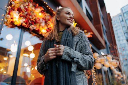 Téléchargez les photos : Jeune femme souriante heureuse marchant dans la rue de la ville décorée par des guirlandes de vacances rougeoyantes sur fond de bâtiments. Vue du bas et du gros plan. - en image libre de droit