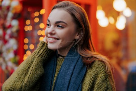 Téléchargez les photos : Portrait de jeune femme souriante heureuse appréciant par vacances guirlandes rougeoyantes sur la rue décorée de la ville. Vue rapprochée. - en image libre de droit