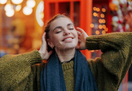 Téléchargez les photos : Portrait de jeune femme souriante heureuse jouissant avec les yeux fermés par des guirlandes rougeoyantes vacances sur la rue décorée de la ville. Vue rapprochée. - en image libre de droit