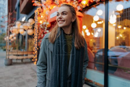 Téléchargez les photos : Jeune femme souriante heureuse marchant dans la rue de la ville décorée par des guirlandes de vacances rougeoyantes sur fond de bâtiments. Vue rapprochée. - en image libre de droit
