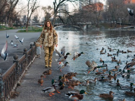 Foto de Mujer soldado ucraniano caminando en el parque durante las vacaciones cerca del lago y los patos.Las mujeres y la guerra en Ucrania.Invasión militar rusa. Traducción del ucraniano: Fuerzas Armadas de Ucrania, apellido. - Imagen libre de derechos