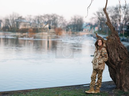 Foto de Mujer soldado ucraniano descansando en el parque durante las vacaciones cerca del lago. Mujeres y guerra en Ucrania. Invasión militar rusa. Traducción del ucraniano: Fuerzas Armadas de Ucrania, apellido. - Imagen libre de derechos