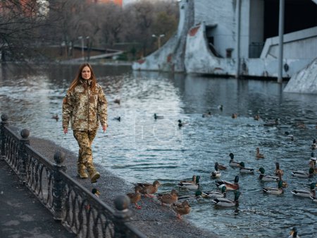 Foto de Mujer soldado ucraniano caminando en el parque durante las vacaciones cerca del lago y los patos.Las mujeres y la guerra en Ucrania.Invasión militar rusa. Traducción del ucraniano: Fuerzas Armadas de Ucrania, apellido. - Imagen libre de derechos