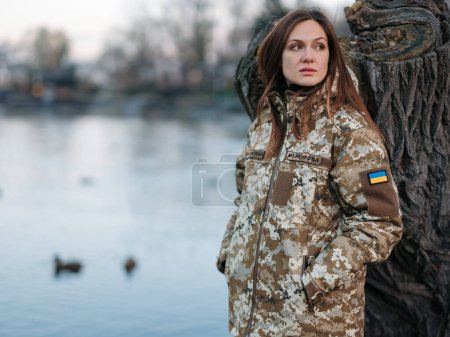 Foto de Mujer soldado ucraniano descansando en el parque durante las vacaciones cerca del lago. Mujeres y guerra en Ucrania. Invasión militar rusa. Traducción del ucraniano: Fuerzas Armadas de Ucrania, apellido. - Imagen libre de derechos