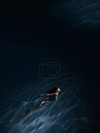 Photo for Underwater shoot of beautiful girl in white dress swimming in water through sunbeams. Ballerina against water surface background with rays of lights. - Royalty Free Image