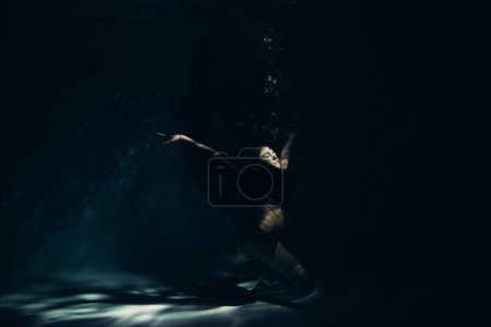 Photo for Underwater shoot of beautiful ballerina in black dress swimming and dancing in water through sunbeams. Ballerina against water background. - Royalty Free Image
