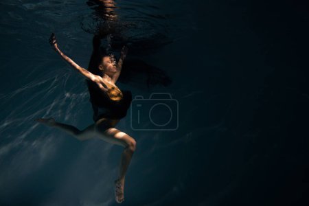 Photo for Underwater shoot of beautiful ballerina in black dress swimming and dancing in water through sunbeams. Ballerina against water background. - Royalty Free Image
