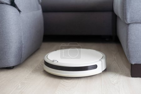 A white robot vacuum cleaner vacuums along the skirting Board near a large window through which the bright sun shines. close-up, beige colors, small depth of field.