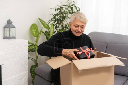 Photo for Beautiful senior woman is holding cardboard box sitting on sofa at home - Royalty Free Image