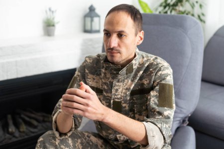 Téléchargez les photos : Thoughtful military man staring aside, holding palms by mouth, sitting on couch at home. Young soldier visiting psychologist, suffering from posttraumatic stress, closeup photo, copy space - en image libre de droit