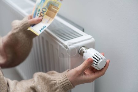 Téléchargez les photos : Femme tenant de l'argent devant le radiateur de chauffage. Paiement pour le chauffage en hiver
. - en image libre de droit