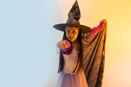 Photo for Happy scary Halloween concept: little witch girl playing paranormal Halloween role and doing scary look on a yellow background. Kid scaring and making Boo in carnival witch costume or wizard costume. - Royalty Free Image
