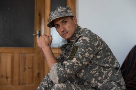 Téléchargez les photos : Homme malheureux en uniforme de camouflage assis au fauteuil sur fond de studio blanc, touchant la tête, soldat souffrant de trouble de stress post-traumatique, rentrant chez lui après le service militaire - en image libre de droit