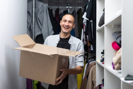Happy cheerful ethnic male in casual clothes carrying cardboard box and looking at camera on moving day with cartons and drawing easels on blurred background