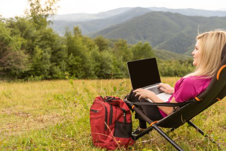 Photo for A woman in a sports warm suit works on a laptop outdoors in a mountainous area - Royalty Free Image