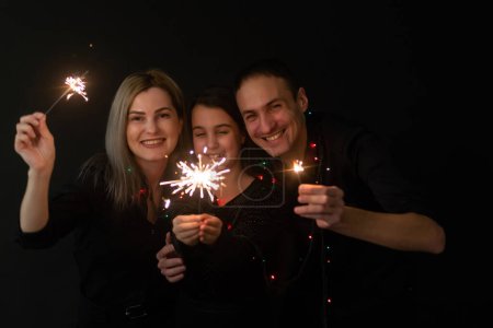Photo for Happy family in black - woman, man and little girl, with a sparkler in hand the New Years Christmas at home. The girl at the father on hands. New year 2023 - Royalty Free Image
