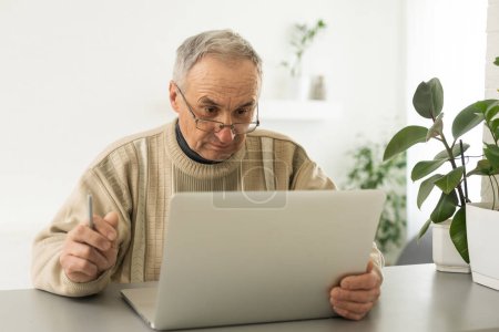 Téléchargez les photos : Bel homme mature dans occasionnel assis dans le bureau à la maison et travaillant à l'ordinateur portable. - en image libre de droit