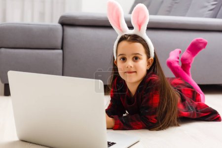 Téléchargez les photos : Petite fille avec son lapin en utilisant l'ordinateur ensemble se préparant pour Pâques- - en image libre de droit