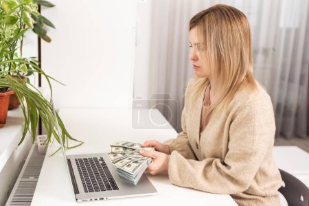 Photo for Businesswoman sitting in her office and holding money near laptop. - Royalty Free Image