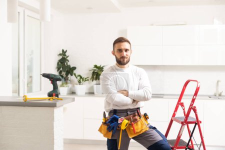 Home repairs. Portrait of professional male repairman with metal ladder. Smiling man in work overalls stands in middle of room in which he has finished making repairs