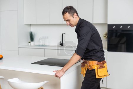 Téléchargez les photos : Homme réparation électrique appartement de surface de cuisine. Installation de plaque de cuisson dans le comptoir. L'homme a acheté de nouveaux meubles et appareils de cuisine. Processus d'installation d'un électricien appareils ménagers - en image libre de droit