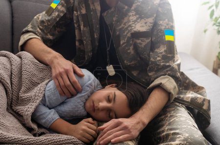 Photo for Soldier in Ukrainian military uniform kissing his daughter while she sleeping on sofa at home. Family reunion - Royalty Free Image
