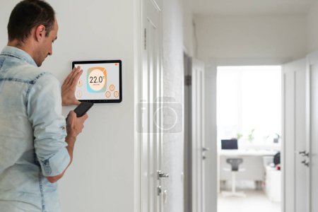 Junger Mann steuert mit Smartphone und digitalem Touchscreen die Temperatur im Wohnzimmer. Konzept der Heizungssteuerung in einem Smart Home. Hochwertiges Foto