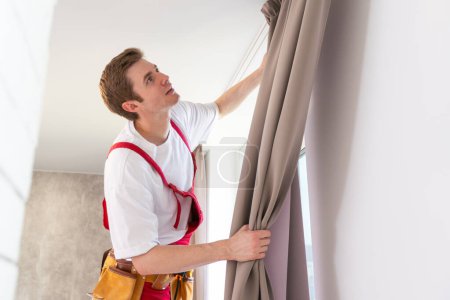 Téléchargez les photos : Un réparateur masculin installe une tringle à rideau et accroche des rideaux dans la maison. services d'aide aux tâches ménagères et aux réparations. - en image libre de droit
