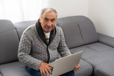 Téléchargez les photos : Homme plus âgé assis sur le canapé, souriant à l'écran d'ordinateur à la maison. Photo de haute qualité - en image libre de droit