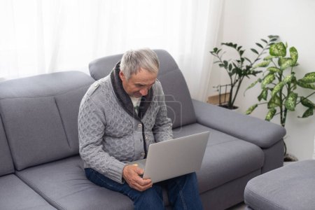 Téléchargez les photos : Homme plus âgé assis sur le canapé, souriant à l'écran d'ordinateur à la maison. Photo de haute qualité - en image libre de droit