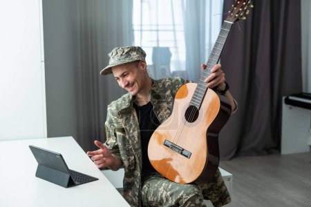 Téléchargez les photos : Joyeux jeune militaire souriant portant l'uniforme kaki tenant la guitare. Photo de haute qualité - en image libre de droit