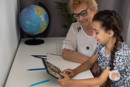 Happy mature grandmother with adorable little granddaughter using tablet at home together, excited middle aged woman and cute kid looking at device screen.