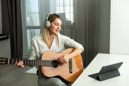 Serene young woman in wireless headphones using acoustic musical instrument while sitting with laptop. High quality photo