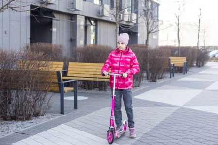 Chica deportiva en edad escolar está montando un scooter en el parque en un paseo en un día soleado. El niño está calurosamente en sombrero. Foto de alta calidad