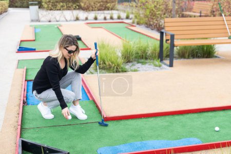 Téléchargez les photos : Femme jouant au mini golf et essayant de mettre la balle dans le trou. Loisirs d'été. Photo de haute qualité - en image libre de droit