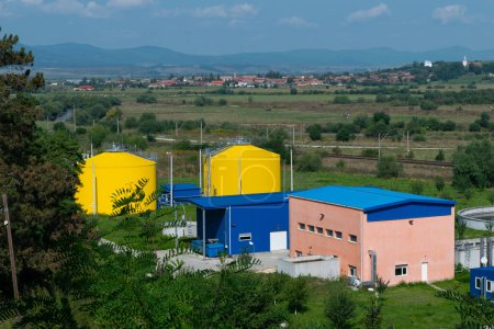 A rural industrial facility showcasing bright yellow tanks, blue structures, and rolling green hills with a distant village