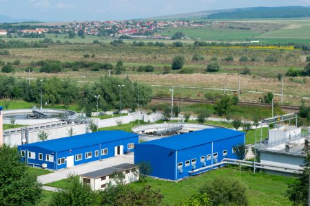 A Industrial Facility with blue Structures and Tanks in Rural Landscape and rolling green hills with a distant village