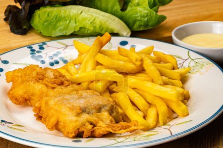 Photo for High angl view of delicious homemade battered fish and french fried in white ceramic dish with a cup of homemade sauce and fresh green vegetable on wooden table. - Royalty Free Image