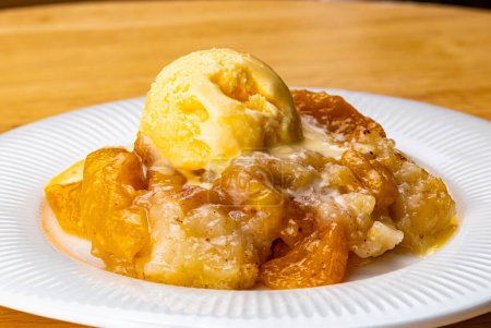 View of juicy homemade delicious peach cobbler with a scoop of vanilla ice cream in white ceramic dish on wooden board.